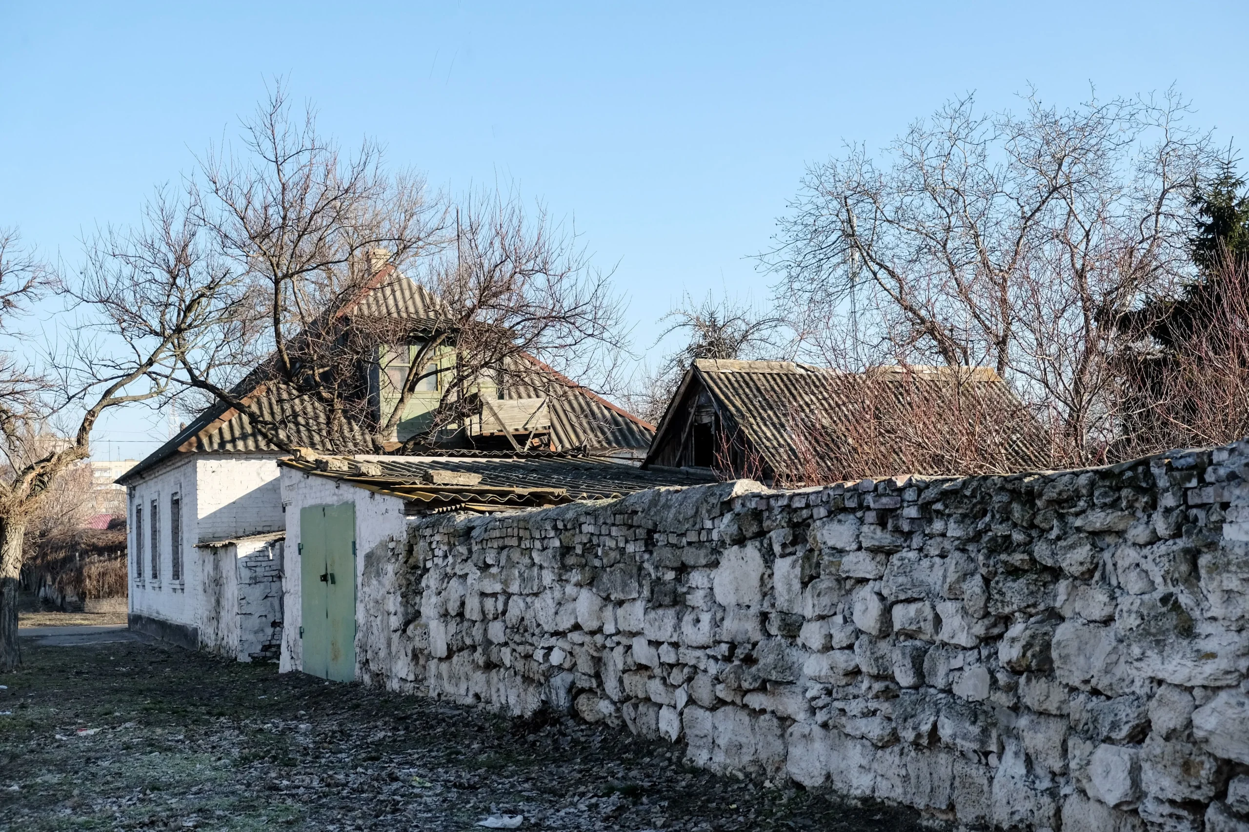 Старий білий будинок з камяним парканом і голими деревами під чистим небом
