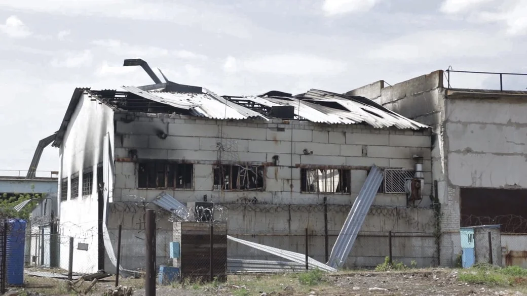 Зруйнована біла будівля з обгорілим дахом і звисаючими металевими листами за колючим дротом