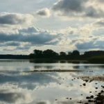 Спокійне озеро віддзеркалює хмари біля лісу та кам’янистого берега