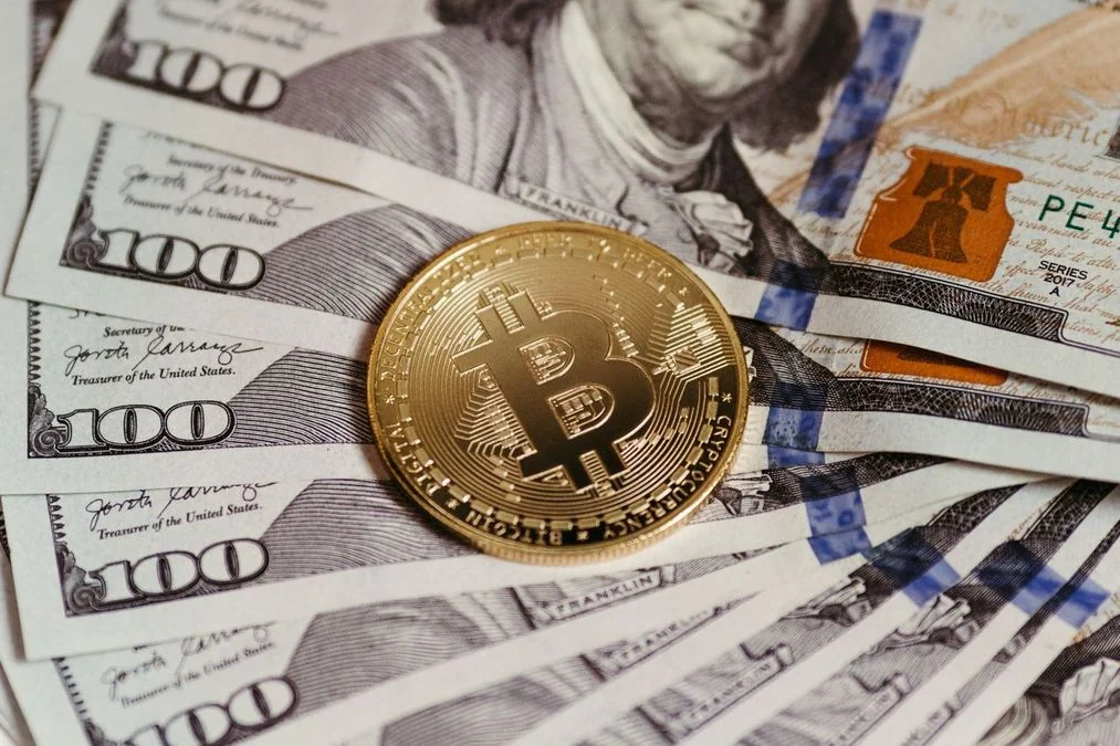 Золота монета Bitcoin лежить на розкладених стодоларових банкнотах